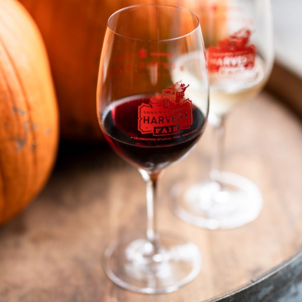 A glass of red wine sits on top of a wine barrel. Pumpkins are slightly visible in the background, as is an empty wine glass.